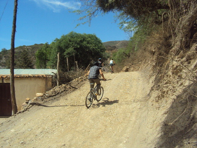 Tres ciclistas de montaña ascendiendo por un camino rural en Yotala junto a una casa tradicional, durante el tour de Bolivian Full Explorer