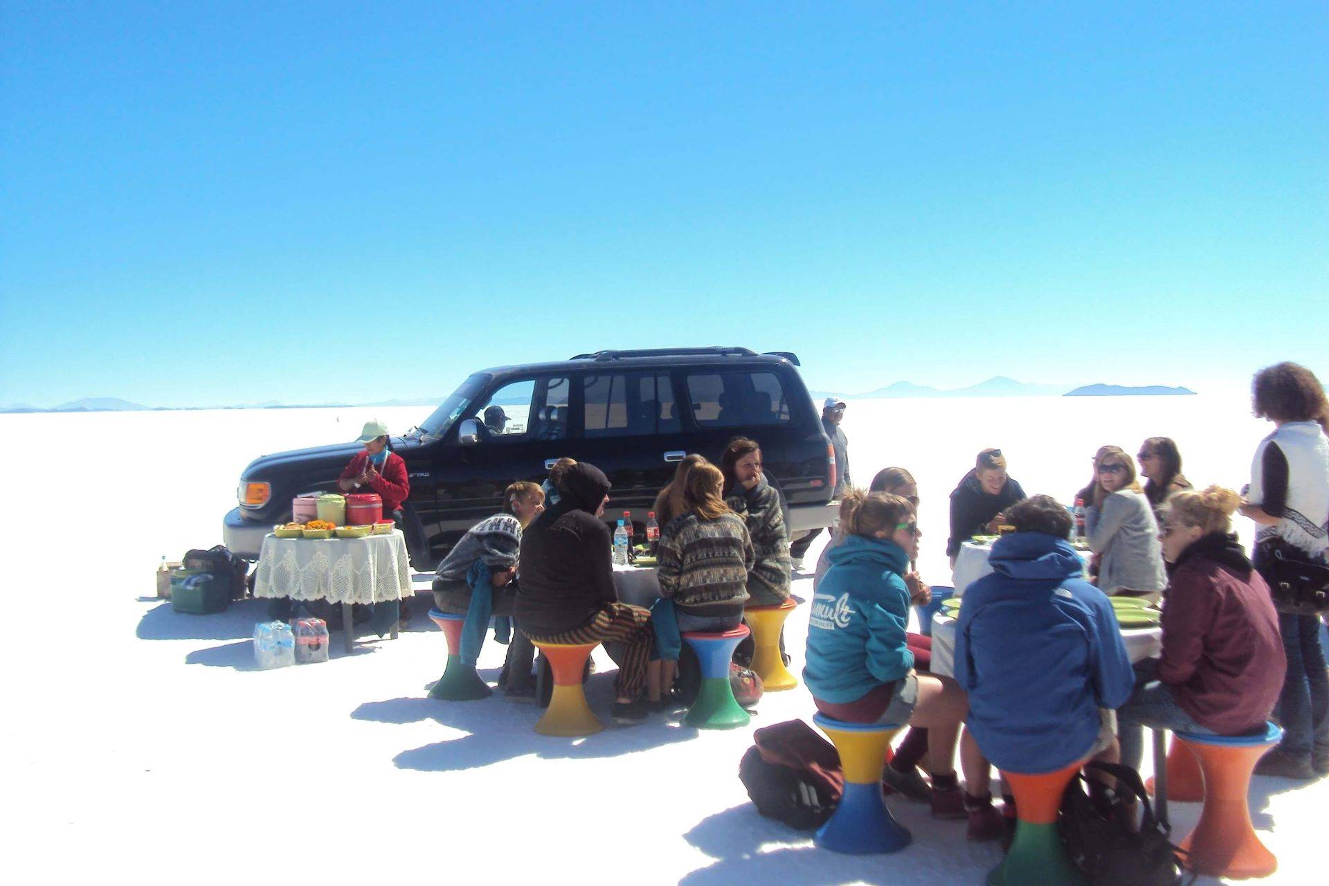 Almuerzo privado en el Salar de Uyuni con Bolivian Full Explorer: Grupo de turistas almorzando sobre el desierto de sal con taburetes de colores y jeep 4x4.