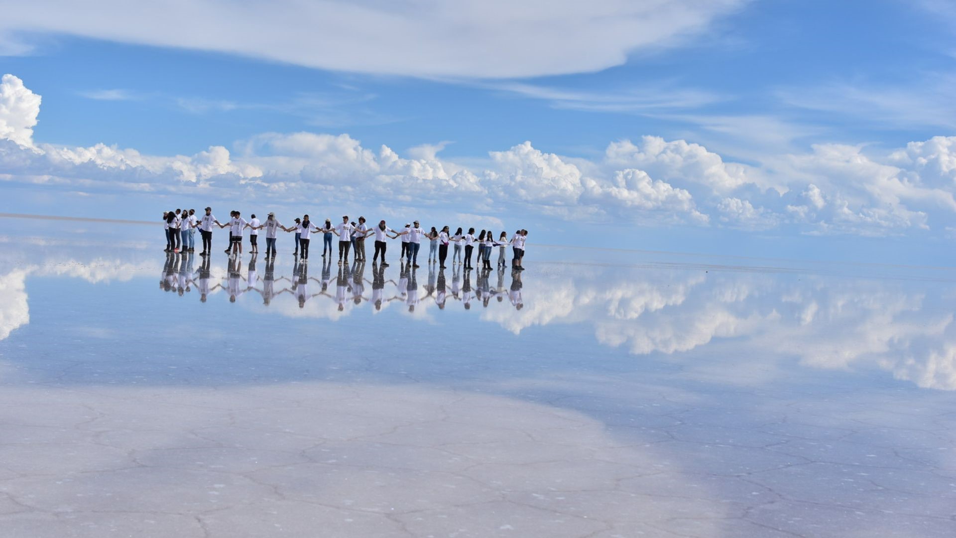 Group of travelers holding hands with a perfect mirror reflection on the Uyuni Salt Flats – Experience by Bolivian Full Explorer.