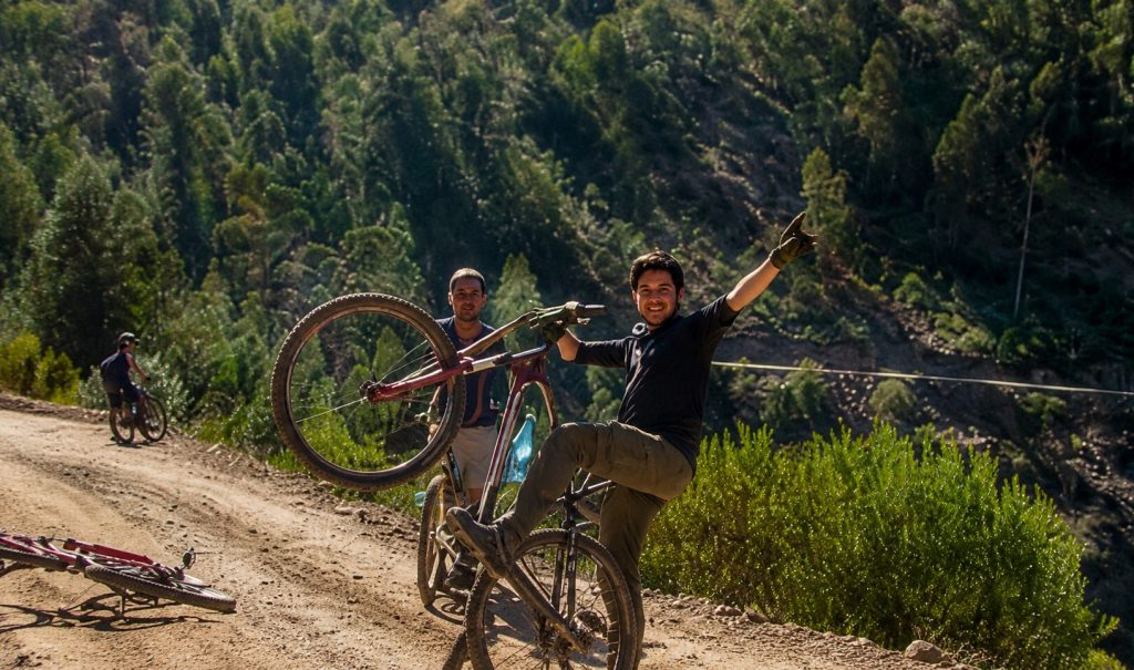 Two cyclists posing with mountain bikes on a dirt trail surrounded by wooded hills.